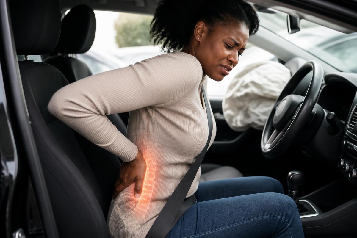 Seat-belted driver supporting her lower back after a collision, with the lumbar spine highlighted